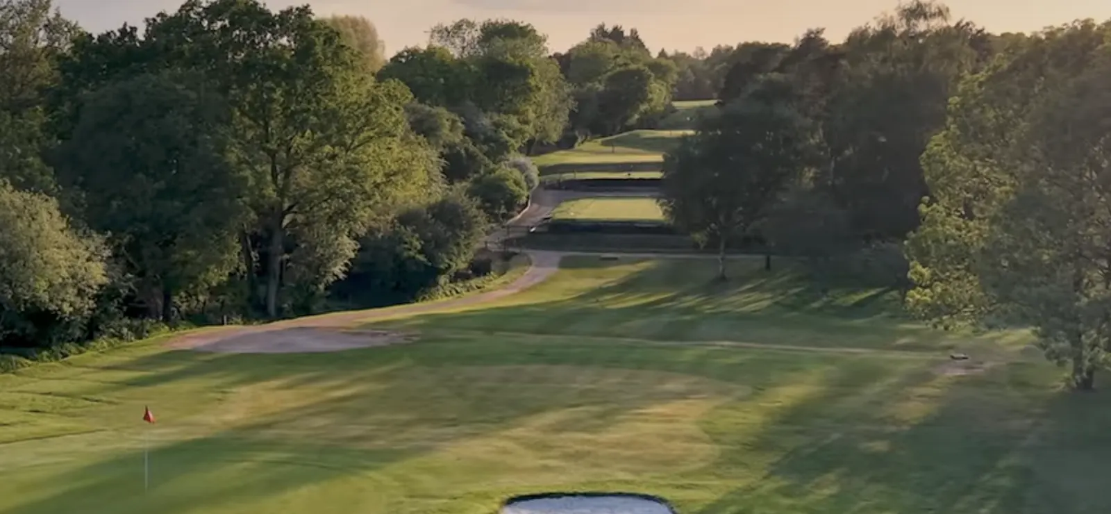 A golf hole seen from above in evening light: a green with the flag in, a bunker below it, and the fairway running away between trees.